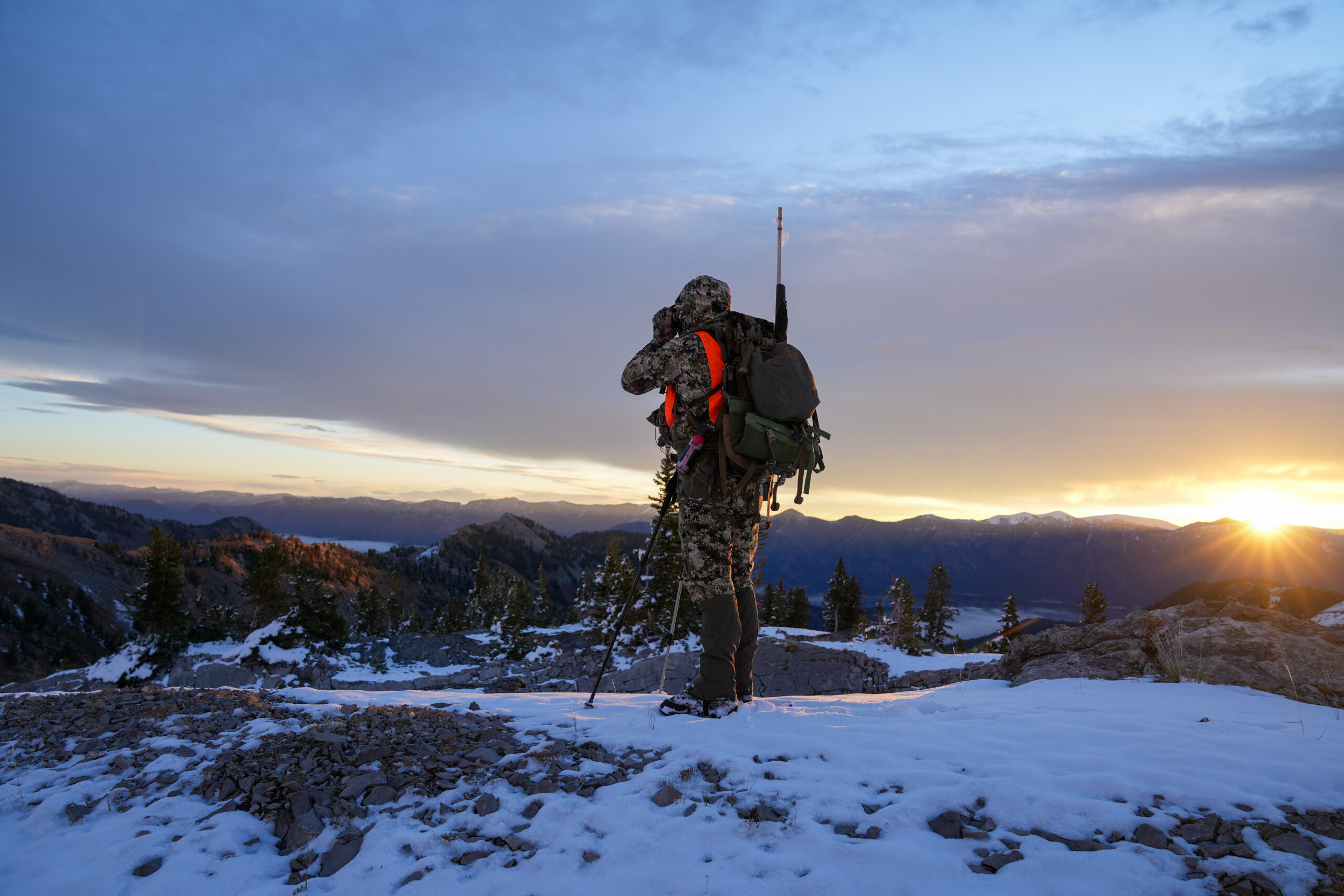 Hunter in camouflage scanning a snowy ridge for deer at sunrise